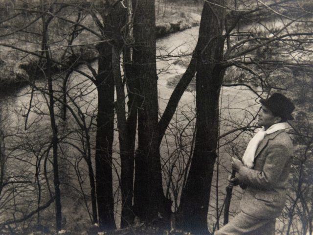 In this black and white photograph, a man leans against a small tree to overlook a river weaving into the distance.