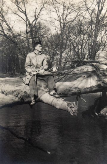 In this vertical black and white photograph, a man sits on a thick tree trunk, fallen over a river.