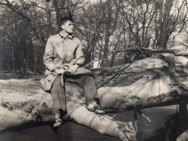 In this vertical black and white photograph, a man sits on a thick tree trunk, fallen over a river.