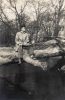In this vertical black and white photograph, a man sits on a thick tree trunk, fallen over a river.