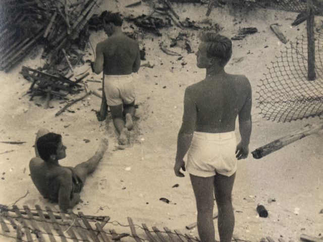 In this image, a man faces away from the camera. He is shirtless and overlooking a beach with wooden walkways.