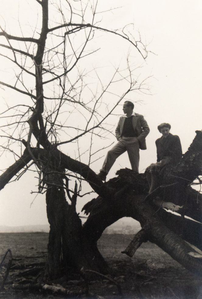 In this black and white photograph, a silhouette of a man stands on a warped tree that reaches towards the earth.
