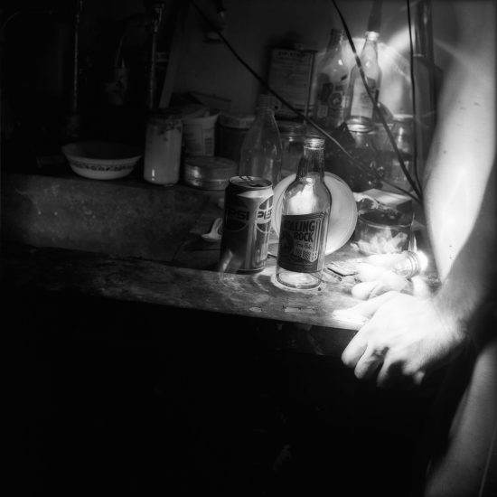This black and white photograph shows a table with a beer bottle. The bright, ghostly hands of a naked man are gripping the table's edge.