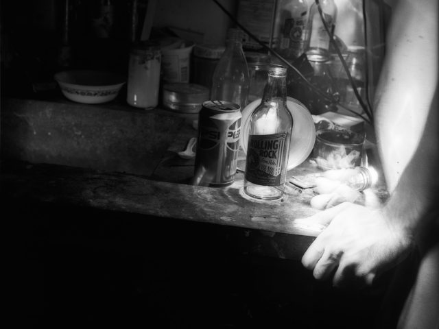 This black and white photograph shows a table with a beer bottle. The bright, ghostly hands of a naked man are gripping the table's edge.