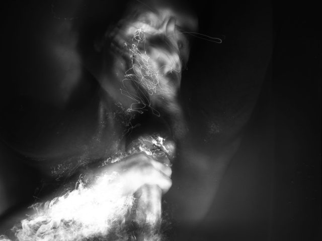 This black and white photograph shows the naked body of a man lying in water. The water reflects the light brightly.