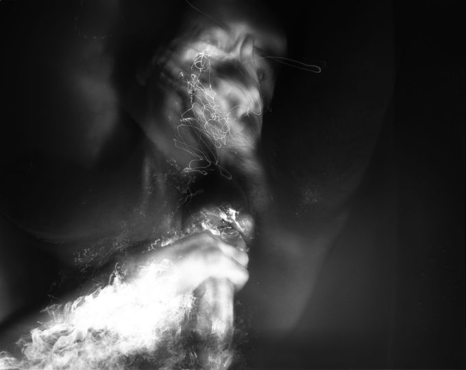 This black and white photograph shows the naked body of a man lying in water. The water reflects the light brightly.
