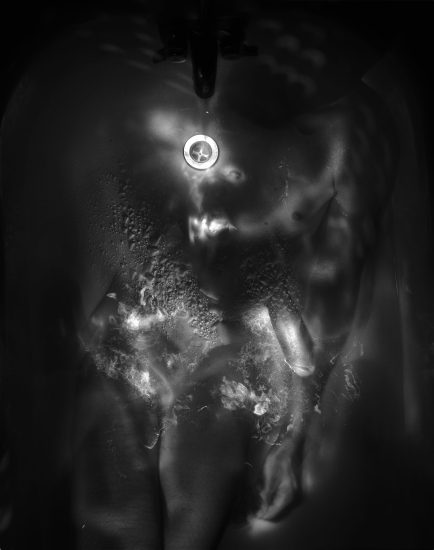 This black and white photograph shows the naked body of a man lying in water that reflects the light.