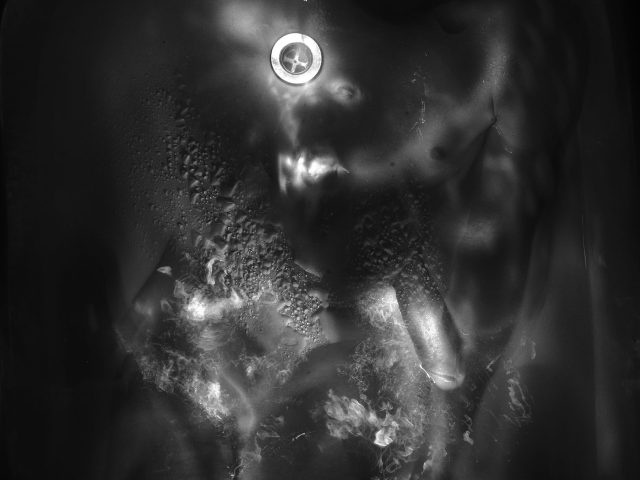 This black and white photograph shows the naked body of a man lying in water that reflects the light.