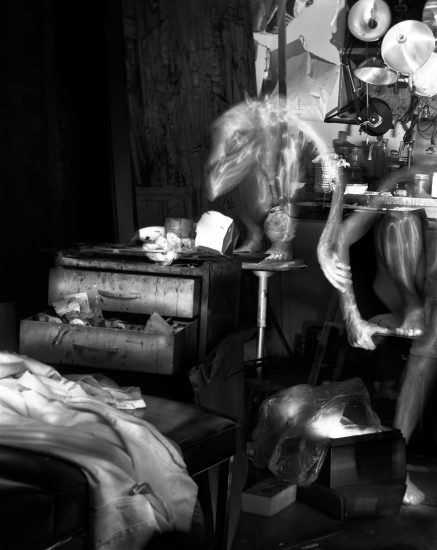 This black and white photograph shows the ghostly figures of naked men in an apartment. They crouch on, and lean against, stools and tables strewn across the crowded room.