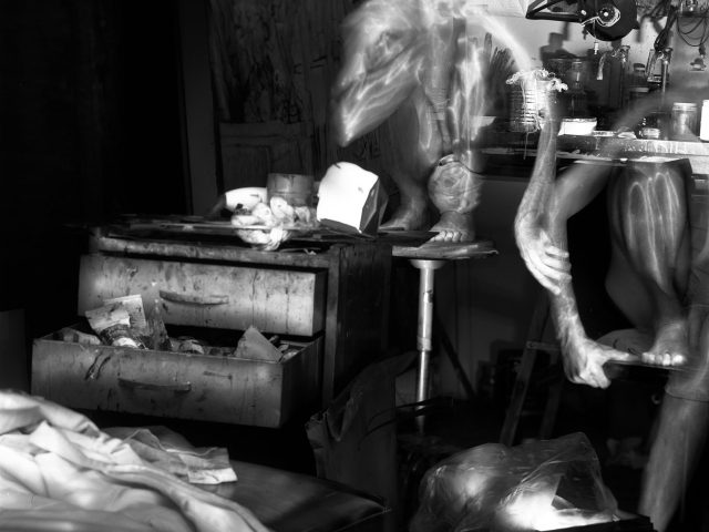 This black and white photograph shows the ghostly figures of naked men in an apartment. They crouch on, and lean against, stools and tables strewn across the crowded room.