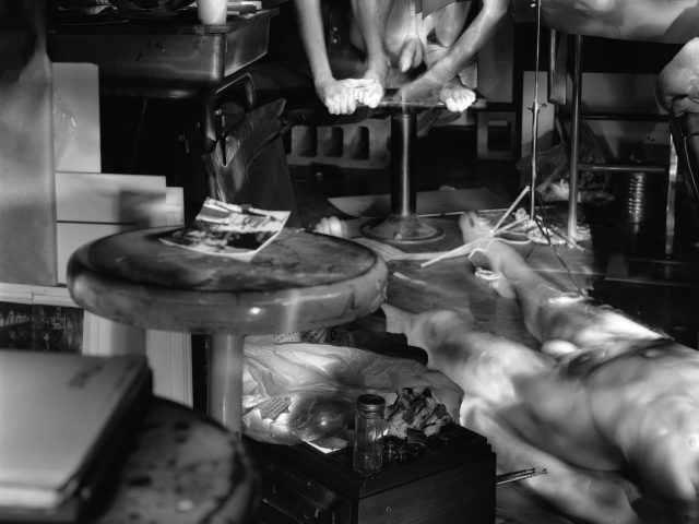 This photograph shows a few naked men, one lying on the ground, and one crouched on a chair. They are seen in the interior of an apartment, covered with various items.