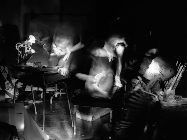 This black and white photograph shows a crowded interior space where a human skeleton leans against a table where a can of paint brushes sits.