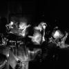 This square black and white photograph shows a mass of bodies in a cramped room, containing odd nick knack such as a small skeleton. The naked bodies are slightly blurred with motion.