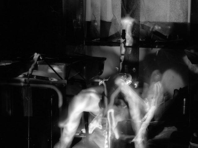 This image shows the ghostly, glowing figures of naked men in an apartment. The curtain in the window behind them is blurred with motion.