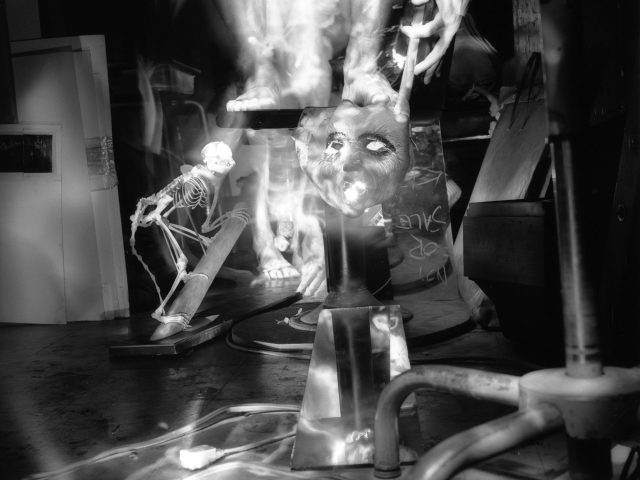 This black and white photograph shows the ghostly figure of a naked man, standing next to the skeleton of a monkey in a crowded apartment. A mask glows in the middle of the frame.