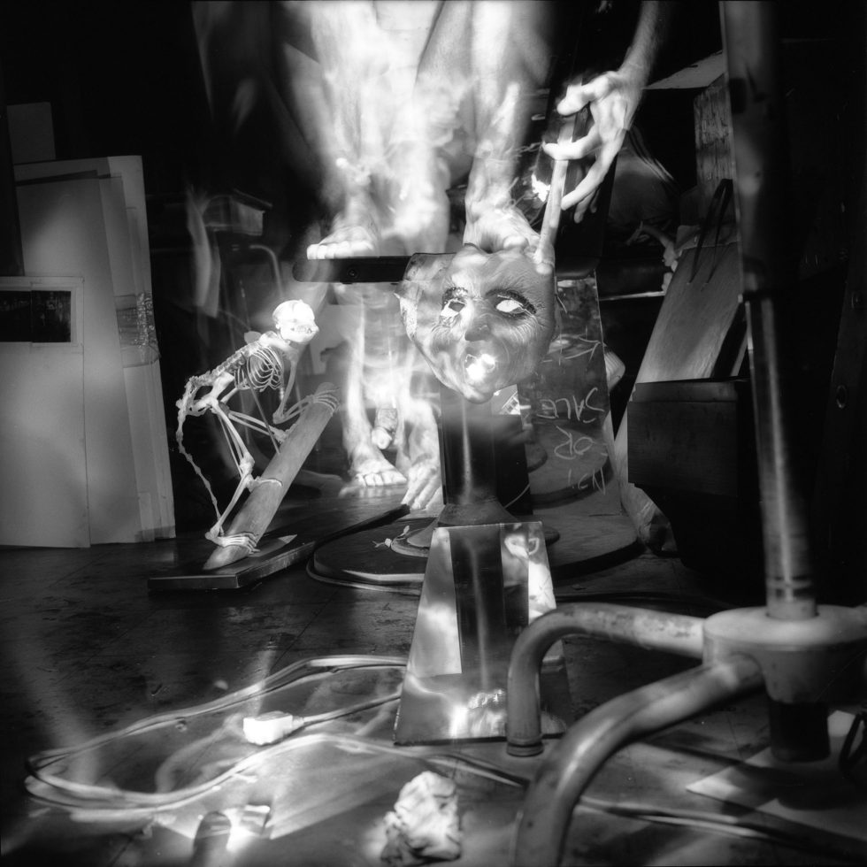 This black and white photograph shows the ghostly figure of a naked man, standing next to the skeleton of a monkey in a crowded apartment. A mask glows in the middle of the frame.