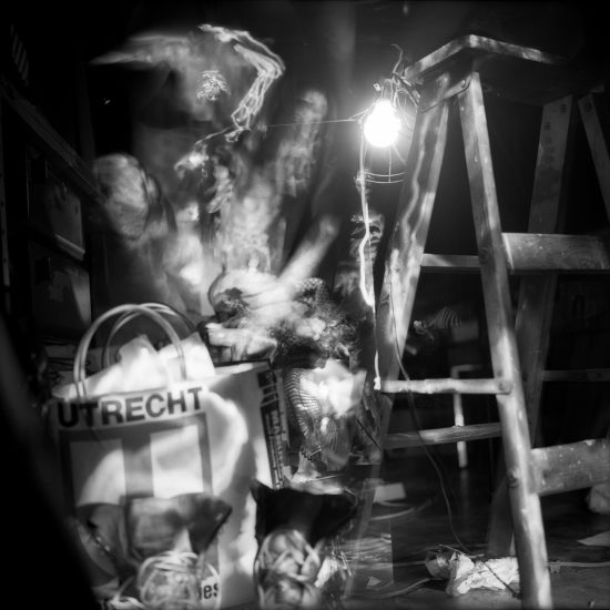 This black and white photograph shows the blurry figure of a naked man standing in a crowded room with a ladder.