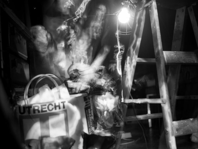 This black and white photograph shows the blurry figure of a naked man standing in a crowded room with a ladder.