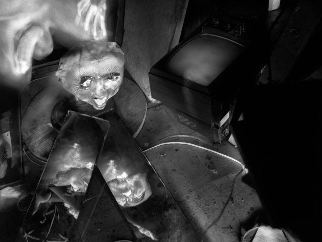 This black and white photograph shows a messy interior space, where a man's ghostly hand brushes the top of a mask placed on top of a table.