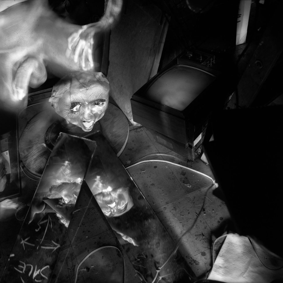 This black and white photograph shows a messy interior space, where a man's ghostly hand brushes the top of a mask placed on top of a table.