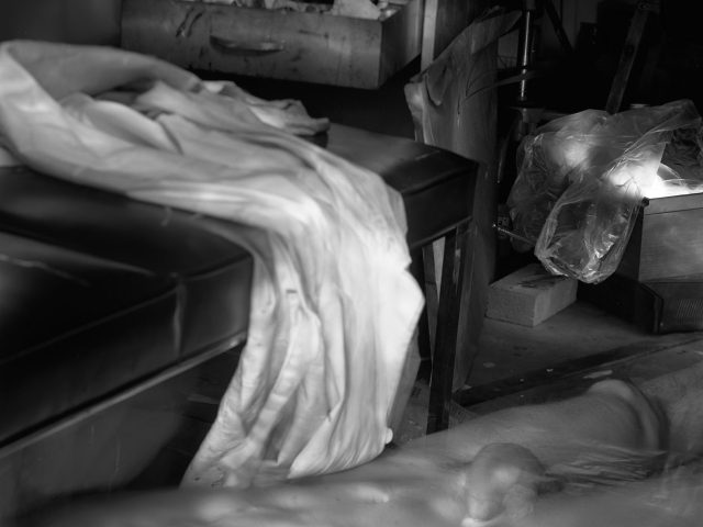 This photograph shows a crowded studio space where a naked man lies on the ground. A long cloth flows down to the ground above him.