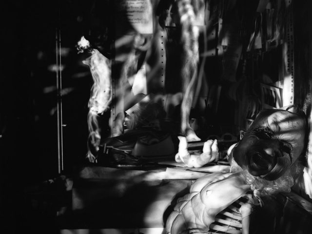 In this black and white photograph, the ghostly figures of naked men stand and sit in tables and chairs strewn across a crowded apartment.