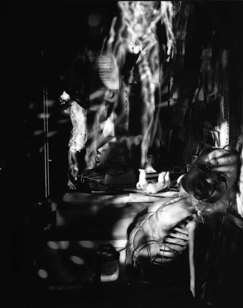 In this black and white photograph, the ghostly figures of naked men stand and sit in tables and chairs strewn across a crowded apartment.