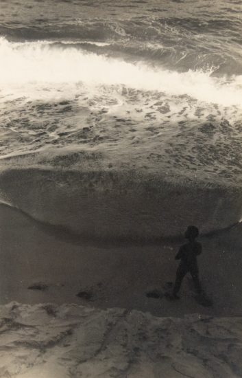 In this faded black and white image, a figure walks down a cloudy beach scape.
