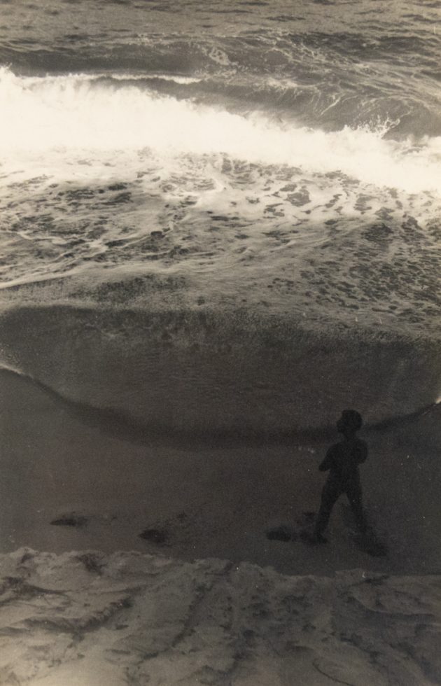 In this faded black and white image, a figure walks down a cloudy beach scape.