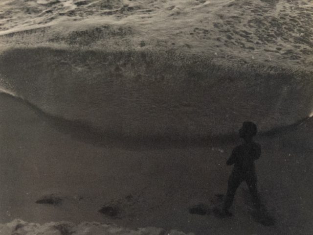 In this faded black and white image, a figure walks down a cloudy beach scape.