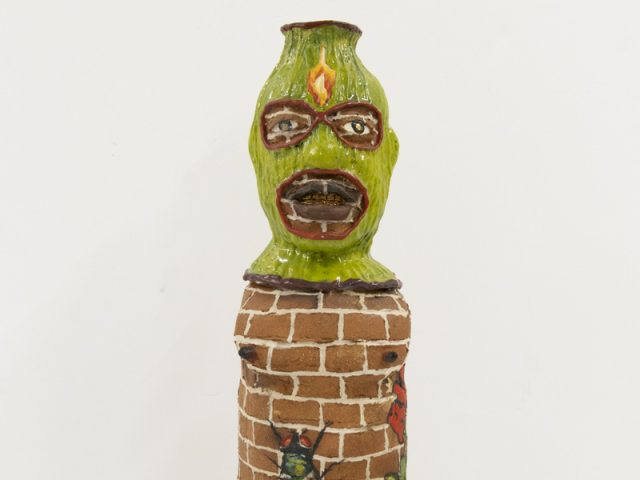 A ceramic vessel depicting a figure with a green hood and a brick body.