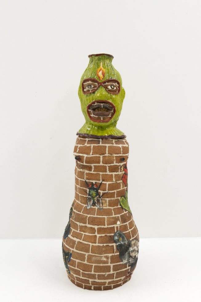 A ceramic vessel depicting a figure with a green hood and a brick body.