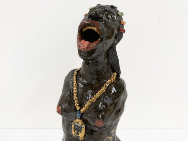 A ceramic vessel with dark brown glazing depicting a figure with no shirt and a gold chain.