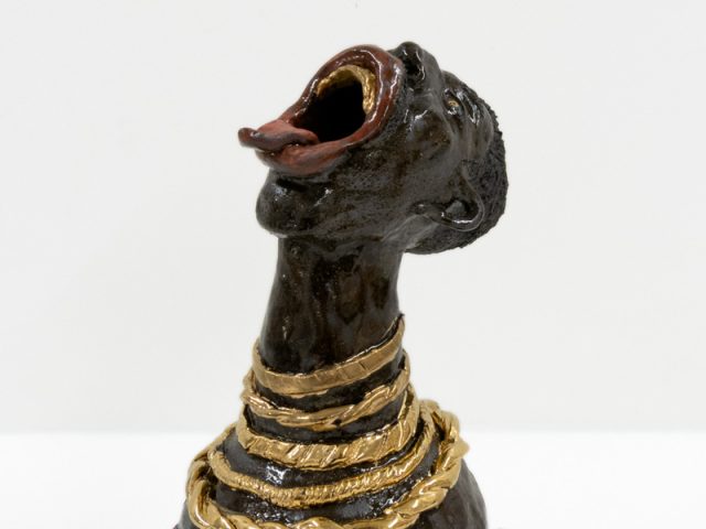 A ceramic vessel with dark brown glazing of a figure with an open mouth and gold necklaces.