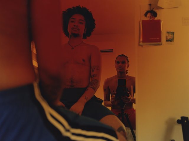 In this color photograph, the photographer and another person sit in front of a mirror. The image is mostly composed of the reflection, where we see them staring into the glass.