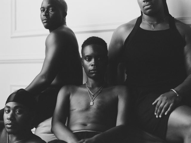 In this black and white photograph, four sitting figures lean against one another and stare at the camera.