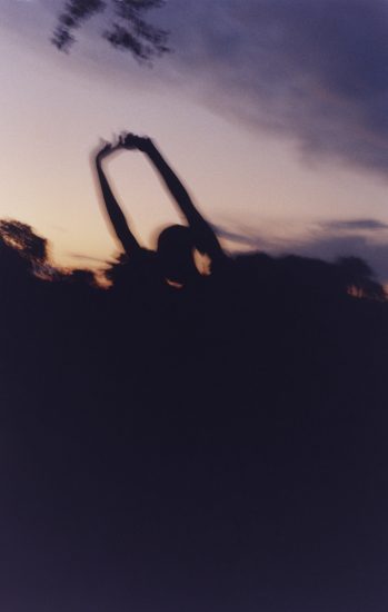 In this color photograph, the silhouette of a figure with their arms raised appears against a purple and orange sunset.