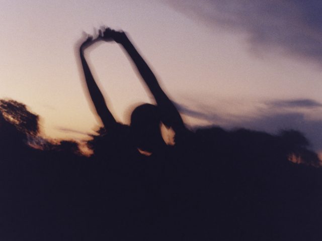 In this color photograph, the silhouette of a figure with their arms raised appears against a purple and orange sunset.