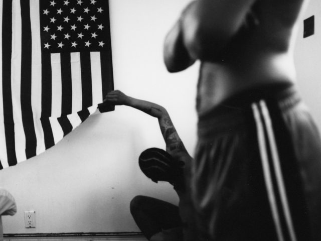 In the foreground of this black and white image, the torso of a figure is seen with folded arms. In the background, another figure squats on the ground and reaches up to lift the corner of a large American flag, hanging from the white wall.