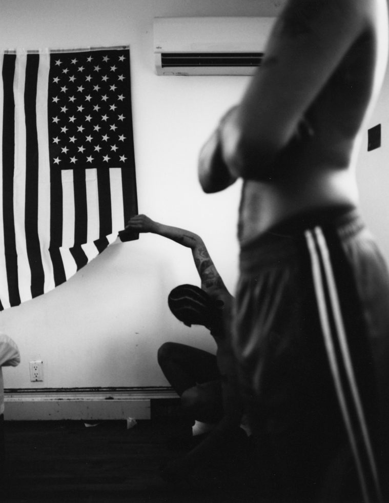 In the foreground of this black and white image, the torso of a figure is seen with folded arms. In the background, another figure squats on the ground and reaches up to lift the corner of a large American flag, hanging from the white wall.