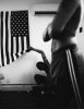 In the foreground of this black and white image, the torso of a figure is seen with folded arms. In the background, another figure squats on the ground and reaches up to lift the corner of a large American flag, hanging from the white wall.