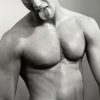 This black and white photograph shows the shirtless body of a man, seen from the waist up. He sticks his tongue out.