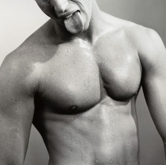 This black and white photograph shows the shirtless body of a man, seen from the waist up. He sticks his tongue out.
