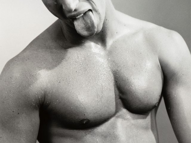 This black and white photograph shows the shirtless body of a man, seen from the waist up. He sticks his tongue out.