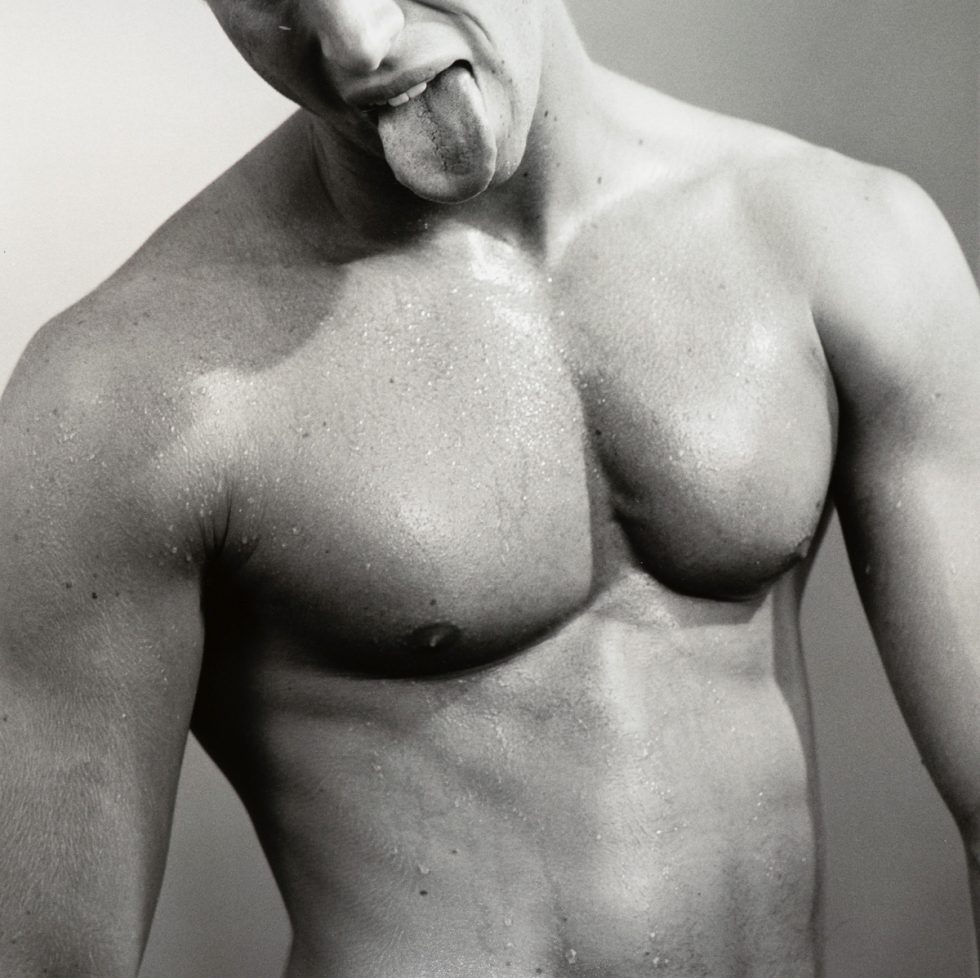 This black and white photograph shows the shirtless body of a man, seen from the waist up. He sticks his tongue out.