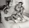 This black and white photograph shows three men lying together on a bed with white sheets. They smile at the camera.