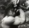 This black and white photograph shows a man flexing his large arm muscles. His arm wraps around his head as he gazes at the camera.
