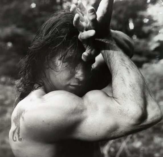 This black and white photograph shows a man flexing his large arm muscles. His arm wraps around his head as he gazes at the camera.