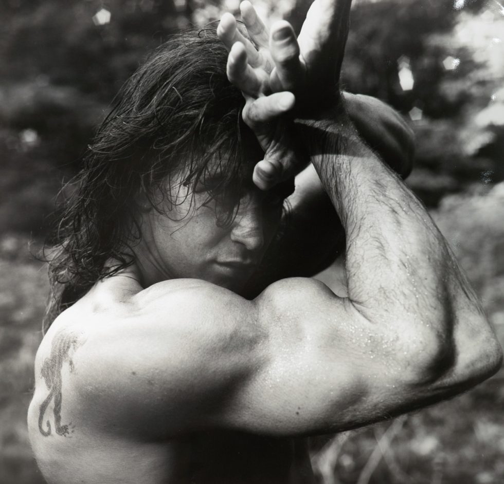 This black and white photograph shows a man flexing his large arm muscles. His arm wraps around his head as he gazes at the camera.