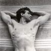 This black and white photograph shows a shirtless man lying on a towel with his arms behind his head.